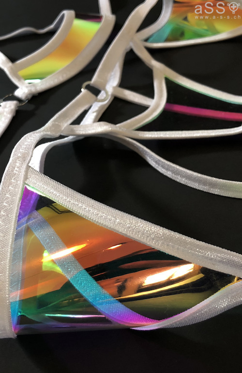 Conjunto de lencería muy inusual en una combinación de tirantes elásticos con vinilo transparente iridiscente. El top es tipo balconet y no cubre los pezones. Las bragas tampoco cubren la zona íntima. Las tiras elásticas bajan desde la braguita hasta los muslos, donde se coloca un liguero. El conjunto también incluye un collar y una visera (protección solar).